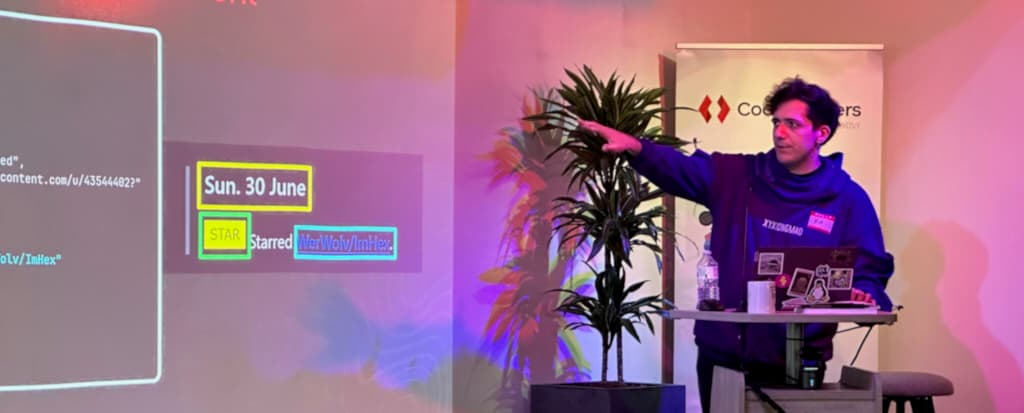 A man wearing a purple hoodie standing at a podium, gesturing toward a projected presentation slide on the wall. The slide displays highlighted text including 'Sun. 30 June', 'STAR', and 'Starred WerWolv/ImHex'. The podium holds a laptop covered in stickers, a clear water bottle, and a white mug. There is a potted plant and a banner with partially visible text in the background, and the room is lit with purple and pink lighting.